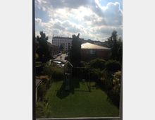 Blick auf einen Garten mit Rasenfläche, einer Schaukel und einer Rutsche, umgeben von Bäumen und Sträuchern. Im Hintergrund sind eine gepflasterte Wohnstraße, mehrere Häuser und ein Kirchturm unter teils bewölktem Himmel zu sehen.