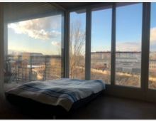 Ein minimalistisches Schlafzimmer mit einem niedrigen Bett vor einer großen Fensterfront, die einen urbanen Ausblick auf Gebäude, Bäume und einen blauen Himmel mit Wolken bietet. Außerhalb der Fenster ist ein Balkon mit einem Geländer sichtbar.