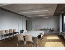 Ein heller, minimalistischer Besprechungsraum mit rechteckigem Tisch und Holzstühlen auf Parkettboden. An einer weißen Wand stehen gestapelte Stühle, und an einer anderen Seite befindet sich ein kleiner Tisch mit Glaskaraffen und Gläsern; große Fenster lassen natürliches Licht herein.