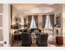 Elegantes Wohnzimmer mit cremefarbenen Wänden, Parkettboden und bodenlangen beige Vorhängen vor großen Fenstern. Es gibt schwarze Polstermöbel, einen Kamin, dekorative Beistelltische und Wandkunst, darunter ein Bild von Mickey Mouse.