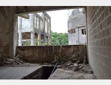 Verfallenes Industriegebäude mit zerbrochenen Wänden, herabgefallenen Trümmern und freigelegtem Beton, das teilweise Blick auf eine weitere Ruine und umliegende Vegetation bietet. Der Raum wirkt verlassen, mit sichtbaren strukturellen Schäden und offenen Fenstern.