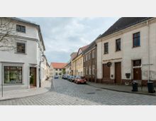 Gepflasterte Straße in einer kleinen Stadt mit zweistöckigen Häusern auf beiden Seiten, darunter Geschäfte wie ein Friseursalon und eine Fleischerei. Die Gebäude haben helle sowie beige Fassaden, rote Ziegeldächer, und am Straßenrand sind geparkte Autos zu sehen.