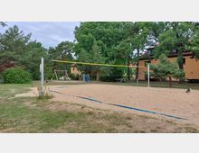Sandplatz mit Volleyballnetz inmitten einer Grünfläche, umgeben von Bäumen und Sträuchern. Im Hintergrund sind ein Spielplatz mit Schaukeln und Rutsche sowie gelbe Gebäude mit roten Dächern zu sehen.