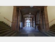 Gewölbter Durchgang in einem Backsteingebäude mit Tonnengewölbe, Rundbogenöffnungen und hängenden Kugelleuchten. Links und rechts führen Treppen mit Holzhandläufen nach oben; am Ende steht eine verglaste Türanlage mit Blick nach draußen.