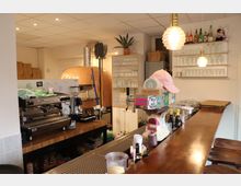 Eine kleine Pizzeria mit Holztheke, Espressomaschine und hölzernem Pizzaofen mit orangefarbener Kachelverkleidung im Hintergrund. Im Raum stehen Regale mit Gläsern und Flaschen, daneben gestapelte Pizzakartons.