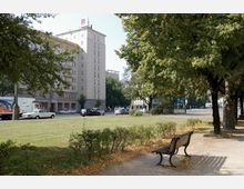 Straßenansicht mit einem mehrstöckigen Wohngebäude im Hintergrund, flankiert von Bäumen und einem kleinen Park mit einer Holzbank im Vordergrund. Autos parken entlang der Straße, und die Szenerie wird von sonnigem Wetter beleuchtet.