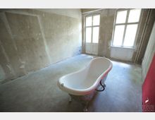 Ein leerer, ungepflegter Raum mit rohen, verputzten Wänden und einem Betonboden. Im Vordergrund steht eine freistehende Badewanne auf dekorativen Füßen, im Hintergrund sind große Fenster und eine Doppeltür, die natürliches Licht hineinlassen.