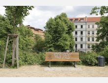 Ein kleiner Parkbereich mit einer Holzbänken, die mit Graffiti besprüht ist, einem jungen Baum mit Stützkonstruktion und einem Mülleimer im Vordergrund. Im Hintergrund sind dichter Baumbestand und Wohnhäuser mit heller und gelber Fassade sichtbar.