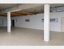 Großer, leerer Innenraum mit hellem Fliesenboden, weißen Wänden und mehreren weißen Stützsäulen. Sichtbare Betondecke mit freiliegenden Holzbalken, Kabeln und Lüftungskanal sowie schmale horizontale Fenster und Türen an der Rückwand.