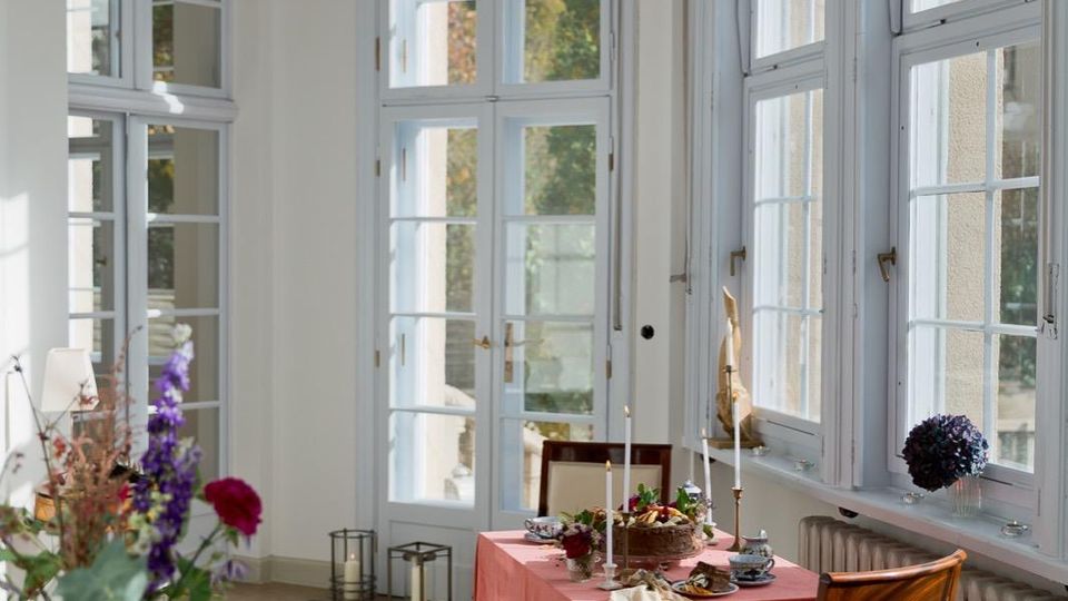 Heller Wintergarten- oder Erkerraum mit hohen, weiß gerahmten Sprossenfenstern an zwei Seiten und hellem Steinboden. In der Mitte steht ein kleiner Tisch mit roter Tischdecke und Kerzen, daneben ein gepolsterter Holzstuhl; entlang der Fenster verläuft ein Heizkörper.