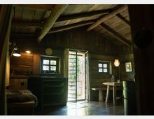 Innenraum einer rustikalen Holzhütte mit schrägem Dach und sichtbaren Holzbalken. Die Ausstattung umfasst kleine Fenster, eine offene Tür mit Blick nach draußen, eine hölzerne Sitzbank und einen Tisch, sowie gedämpfte Beleuchtung durch Lampen.