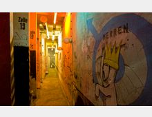 Schmaler Flur mit rauer, bemalter Wand, graffitibedeckten Säulen und orange sowie gelb beleuchteten Lampen. Auf der rechten Wand ist eine große Zeichnung mit einem Kreis und einer Figur mit gelber Krone zu sehen; Beschriftungen wie „Zelle 13“ sind auf den Säulen angebracht.