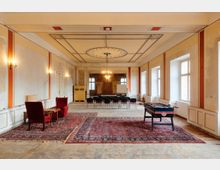 Großer, rechteckiger Saal in einem älteren Gebäude mit hohen Decken, Stuckornamenten und Kronleuchter sowie hohen Fenstern entlang einer Wand. Im Raum stehen Sitzreihen, Teppiche, zwei Sessel mit Beistelltisch und ein Tischkicker; die Wände sind teilweise abgeblättert und renovierungsbedürftig.