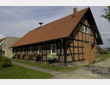 Fachwerkhaus mit Backsteinfassade und roten Dachziegeln in einer ländlichen Umgebung, mit mehreren Fenstern, die Blumenkästen mit bunten Blumen tragen. Das Gebäude steht auf einem Grundstück mit einer Wiese und einem gepflasterten Weg.