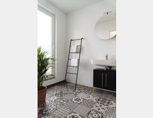 Modernes Badezimmer mit schwarz-weißen gemusterten Fliesen auf dem Boden, einer weißen Wand, einem runden Spiegel über einem Waschbecken mit schwarzem Unterschrank, einer dekorativen Handtuchleiter und einer Pflanze in einem Korb. Links befindet sich ein großes Fenster mit Tageslichteinfall.