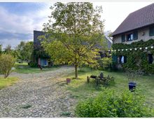 Gepflegter Hofbereich mit Kopfsteinpflasterwegen, umgeben von einem Garten mit Bäumen und Sträuchern. Auf der rechten Seite steht ein mit Efeu bewachsenes Haus mit grünen Fensterläden, links eine Scheune mit blauen Türen; im Vordergrund ein alter Holzwagen und Gartenmöbel.