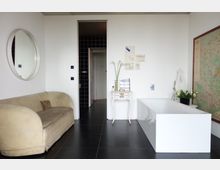 Minimalistisch eingerichteter Raum mit schwarzem Fliesenboden, einem beigen Sofa, einer freistehenden weißen Badewanne und einem kleinen weißen Tisch. An der weißen Wand hängen Zeichnungen und ein Rundspiegel, während auf der rechten Seite eine große Landkarte sichtbar ist.