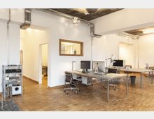 Modernes Büro mit minimalistischem Design, ausgestattet mit großen Schreibtischen, Bürostühlen und Computern. Der Raum hat einen hellen Holzboden, weiße Wände, sichtbare Kabelkanäle an der Decke und eine Trennwand mit Holzrahmenfenster.