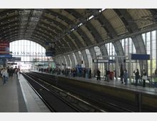 Bahnhofshalle mit geschwungenem Glasdach und Metallstrukturen, zwei Bahnsteige mit wartenden Menschen und Gleisen in der Mitte, umgeben von Werbung, Hinweisschildern und Tageslicht aus großen Fenstern.