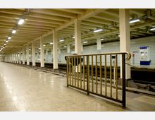 U-Bahn-Station mit einem offenen Bahnsteig, flankiert von weißen Wänden und einer Decke mit sichtbaren Querbalken. Im Vordergrund ein Geländer, im Hintergrund Fahrkartenautomaten und Hinweisbeschilderung.