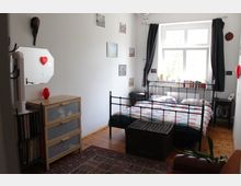 Bright bedroom with a black metal bed frame and patterned bedding, set on a wooden floor with a small rug and a wicker storage bench at the foot of the bed. A large window with dark curtains lights the room, with books and small plants on the sill, and storage drawers and shelves along the left wall.