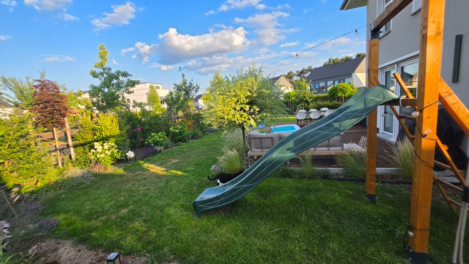 Blick in einen gepflegten Wohnhausgarten mit Rasenfläche, Beeten und mehreren Bäumen; rechts steht eine Holzterrasse am Haus mit Sitzgruppe und kleinem Pool. Im Vordergrund führt eine grüne Rutsche von einer Holzspielkonstruktion in den Garten, darüber sind Lichterketten gespannt.