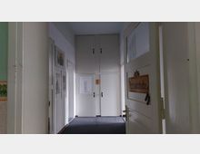 Ein schmaler, heller Flur mit weißen Wänden und dunklem Teppichboden. Auf der rechten Seite ist eine geöffnete Tür mit einem Schild "Heimatstube", während an der linken Wand gerahmte Bilder hängen; am Ende des Flurs befinden sich geschlossene Schranktüren.