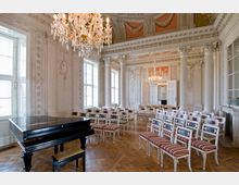 Eleganter Konzertsaal mit verzierten weißen Säulen, einem kunstvollen Kronleuchter, einem schwarzen Flügel im Vordergrund und mehreren Stuhlreihen mit rot-weiß gestreiften Polstern auf einem Parkettboden. Die Wände sind mit Reliefs und Stuckarbeiten geschmückt, und hohe Fenster lassen Tageslicht herein.