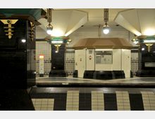 U-Bahn-Station mit Art-déco-Elementen, gekennzeichnet durch schwarz-glänzende Säulen mit goldfarbenen Verzierungen und grünen Akzenten. Im Zentrum steht ein weißes Kontrollhaus mit goldbraunem Dach, umgeben von gefliesten Wänden und einer stilisierten Gitterumzäunung.