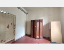 Ein schlichter Innenraum mit abgenutzter Tür mit abblätternder Farbe, einem braunen Holzspind in der Mitte und mehreren flach gestapelten Holzelementen an der rechten Wand. Der Boden ist rötlich gefärbt, und die Wände sind schlicht und hell gehalten.