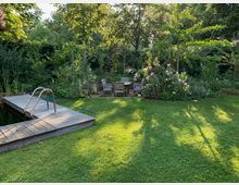 Ein Garten mit einem Holzdeck und Einstiegsmöglichkeit in ein kleines Gewässer auf der linken Seite. Im Hintergrund befinden sich eine Sitzgruppe aus Holzstühlen und ein Tisch, umgeben von Blumenbeeten und dichter Vegetation aus Büschen und Bäumen.