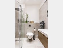 Modernes Badezimmer mit begehbarer Glasdusche links, wandhängendem WC in der Mitte und Waschtisch mit Holzfront rechts. Helle Steinfliesen, ein schwarzer Handtuchheizkörper und eine Ablage mit grünen Glasvasen und Pflegeflaschen prägen den Raum.