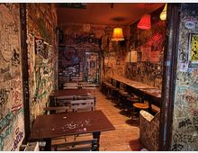 Schmaler Innenraum einer Bar oder eines Cafés mit Holzdielen, langen Holztischen und Hockern sowie mehreren Hängelampen mit farbigen Stoffschirmen. Wände und Türflächen sind dicht mit Graffiti, Stickern und Plakaten bedeckt, wodurch ein stark urbaner, dekorierter Look entsteht.