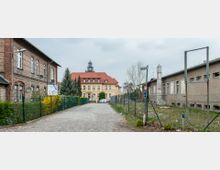 Straßenszene mit einem gepflasterten Weg, flankiert von einem Ziegelgebäude links und einem grauen, verfallenen Gebäude rechts. Im Hintergrund ein historisches gelbes Gebäude mit rotem Dach und einem Glockenturm, umgeben von Bäumen und geparkten Autos.