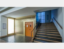 Flur in einem Gebäude mit einer Treppe aus Stein und einem orangen Aufzug. Neben dem Aufzug befindet sich ein beleuchtetes Verzeichnis der Stockwerke, im Hintergrund ein großes Fenster mit Rahmen aus Metall.
