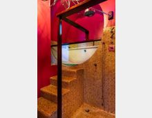 Eine Dusche mit runden, goldfarbenen Mosaikfliesen und einer markanten roten Wand im Hintergrund. Die Dusche verfügt über eine Regendusche, integrierte Armaturen und eine Treppe, die zu einer erhöhten Badewanne hinter einer Glaswand führt.