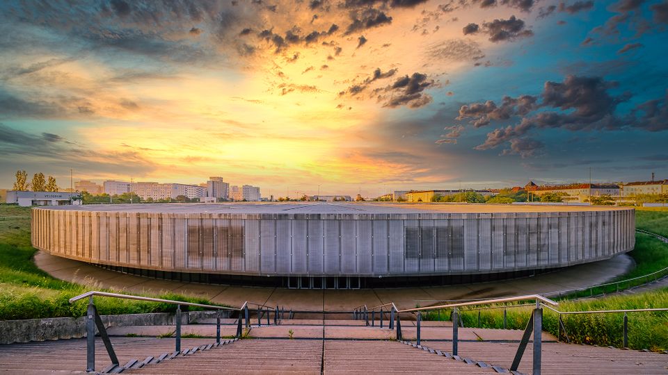 Rundes, modernes Stadiongebäude mit umlaufender Lamellenfassade in flacher Umgebung, davor breite Treppenanlagen mit Geländern und grasbewachsenen Böschungen. Im Hintergrund sind niedrige Stadtgebäude und ein dramatischer, farbiger Abendhimmel mit Wolken zu sehen.