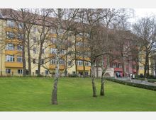 Wohnhäuser in einer parkähnlichen Umgebung mit grünen Rasenflächen und kahlen Bäumen im Vordergrund. Eine Gruppe von Menschen steht vor einem Gebäude mit gelben und roten Fassaden sowie zahlreichen Fenstern.