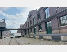 Ziegelgebäude einer alten Lagerhalle mit der Aufschrift „Lagerhalle 4“, großen grünen Türen und Fenstern, umgeben von Gleisen und gepflastertem Boden. Im Vordergrund steht ein weißer Lieferwagen, daneben ein historischer Hafenkran und eine schmale Brücke.