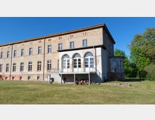 Zweigeschossiges, rechteckiges Gebäude mit verputzter Fassade und sichtbarem Backsteingesims. Eine Terrasse mit Rundbogenfenstern und Metallgeländer ragt hervor, umgeben von einer Rasenfläche und Bäumen im Hintergrund.