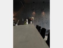 Eine moderne Bar mit einer strukturierten, dunklen Wand und hängenden, schlanken Lampen. Auf der Theke stehen ein Blumenstrauß in einer Vase und dekorative Elemente; schwarze Barhocker sind im Vordergrund sichtbar.