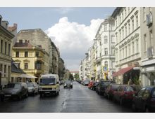 Städtische Straßenansicht mit mehrstöckigen Wohnhäusern auf beiden Seiten, die teils historische Fassadendekorationen aufweisen. Parkende Autos und ein Kleinbus säumen die nasse Straße, während ein U-Bahn-Schild auf eine nahegelegene Station hinweist.