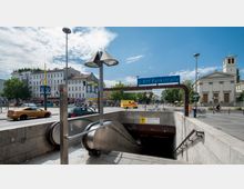 Eingang zur U-Bahn-Station „Pankstraße“ mit Rolltreppe und blauem Stationsschild. Im Hintergrund sind Wohnhäuser, ein gelbes Wandbild, eine Kirche mit Säulenfassade sowie parkende und fahrende Fahrzeuge auf einer belebten Straße zu sehen.