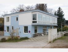 Zweistöckiges Gebäude mit blauer und weißer Fassade, großen Fenstern im Obergeschoss und flachem Dach. Vor dem Gebäude befinden sich ein gepflasterter Weg, Büsche sowie eine kleine Rampe mit Geländer und ein weißes Tor.