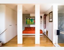 Modernes, helles Zimmer mit Holzboden, einer eingebauten weißen Schrankwand links und einem orangefarbenen Tagesbett in einer Nische, dekoriert mit einem bunten Poster an der Wand. Rechts ist ein schmaler Flur mit gerahmten Bildern und einer Wandhalterung zu sehen.