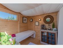 Holzhausinnenraum mit heller Holzverkleidung, einer Behandlungsliege, dekorativen Bildern und einem runden Spiegel an der Wand. Ein kleines Regal mit Glastüren, ein Ventilator und Pflanzenakzente ergänzen die schlichte Einrichtung.