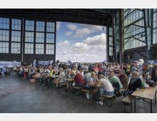Menschenmenge in einer großen, offenen Halle mit hohen Glastüren und Stahlstrukturen. Im Vordergrund sitzen viele Personen an Holzbänken und -tischen, während im Hintergrund weitere Menschen stehen; durch die offene Tür ist ein bewölkter Himmel sichtbar.
