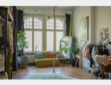 Bright studio-style room with tall arched grid windows, wooden floors, and a central dance pole, decorated with hanging globe lights. Clothing racks line the sides, with a small sofa, large potted plants, a full-length mirror, and an easel near the window.