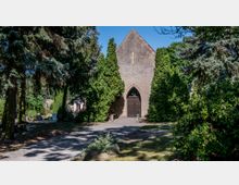 Eine kleine, steinerne Kapelle mit spitzbogigem Eingang, brauner Tür und goldfarbenem Kreuz auf dem Giebel, umgeben von hohen Nadelbäumen und dichtem Grün in einem friedlichen, parkähnlichen Umfeld.
