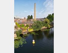 Ehemaliges Industrieareal am Ufer eines Kanals mit Backsteingebäuden und einem hohen, schlanken Schornstein im Hintergrund. Im Vordergrund fließt das Wasser mit Seerosen, Uferbäumen und einer gelb-weißen Boje.
